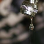 Rubinetto che perde gocce in un ambiente buio, evidenziando il problema del consumo d'acqua e l'aumento della bolletta.